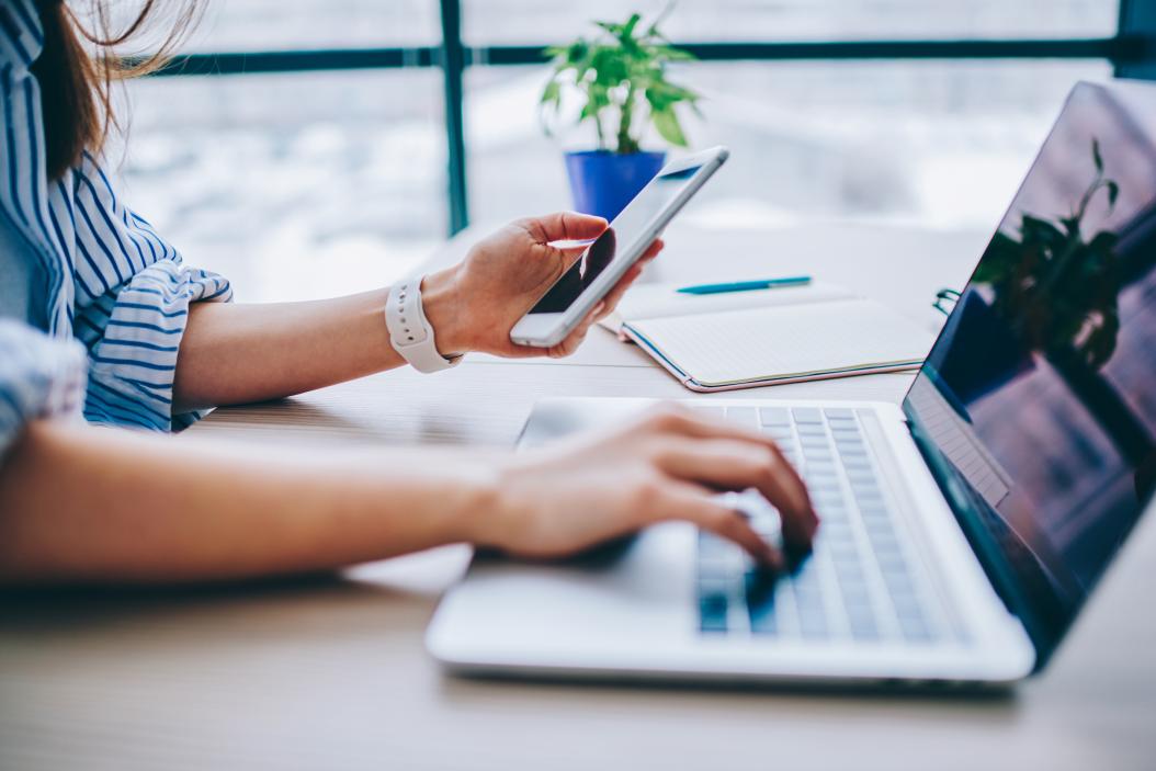 Nahaufnahme einer Frau, die ein Smartphone hält und parallel auf ihrem Laptop tippt