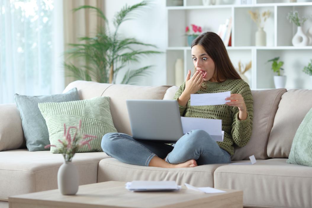 Eine Frau sitzt auf dem Sofa mit dem Laptop auf ihrem Schoß und guckt dabei positiv überrascht.