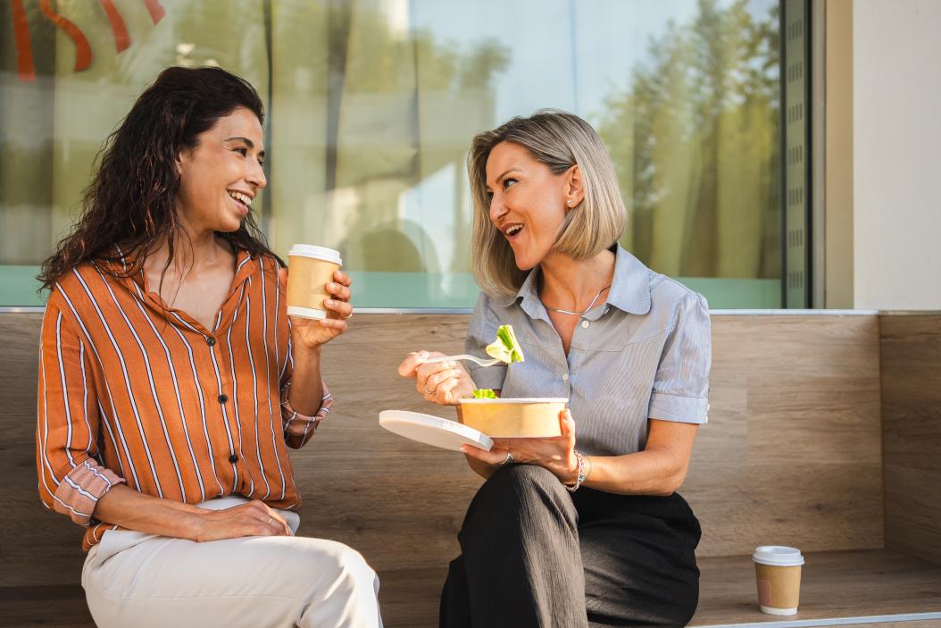 Zwei Geschäftsfrauen beim freundschaftlichen Mittagessen im Freien. Sie essen Salat und trinken Kaffee.