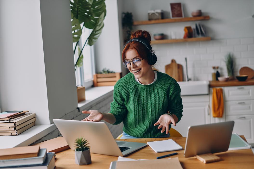 Fröhliche junge Frau mit Kopfhörern nimmt während der Arbeit im Homeoffice an einer Webkonferenz teil.