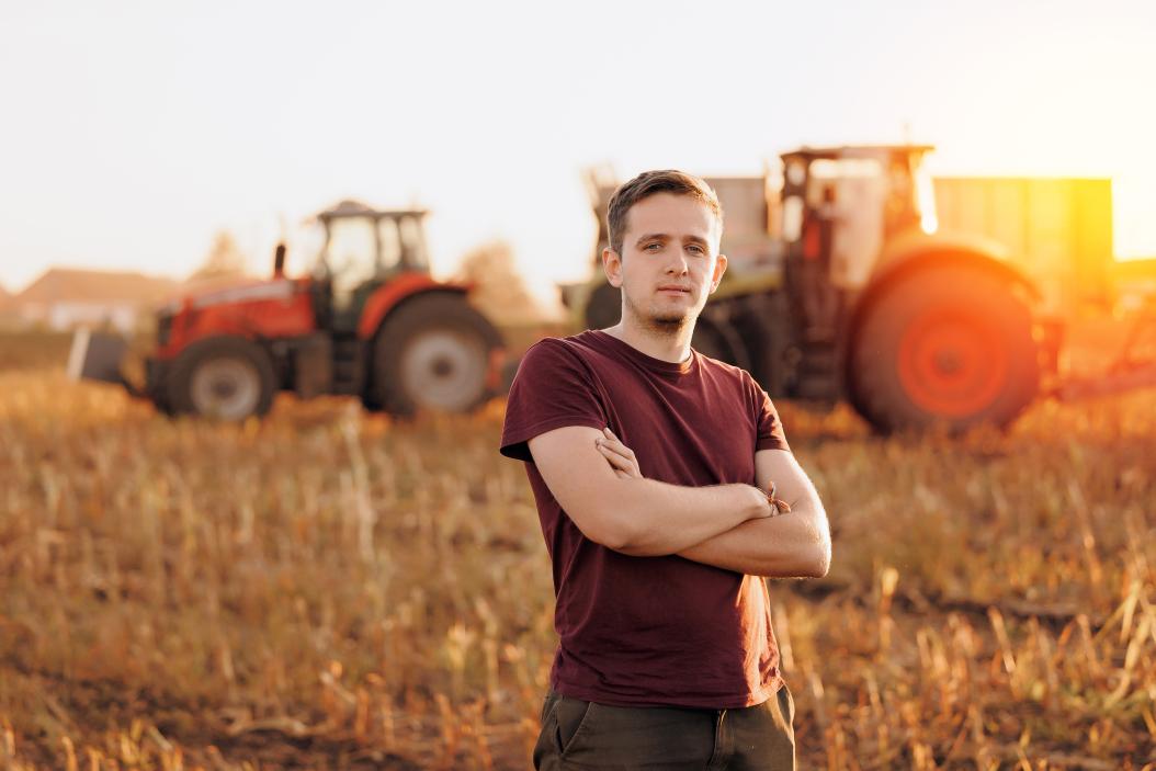 Porträt eines jungen Landwirts in einem Maisfeld, wo im Hintergrund ein Mähdrescher und ein Traktor auf dem abgeernteten Maisfeld zu sehen sind