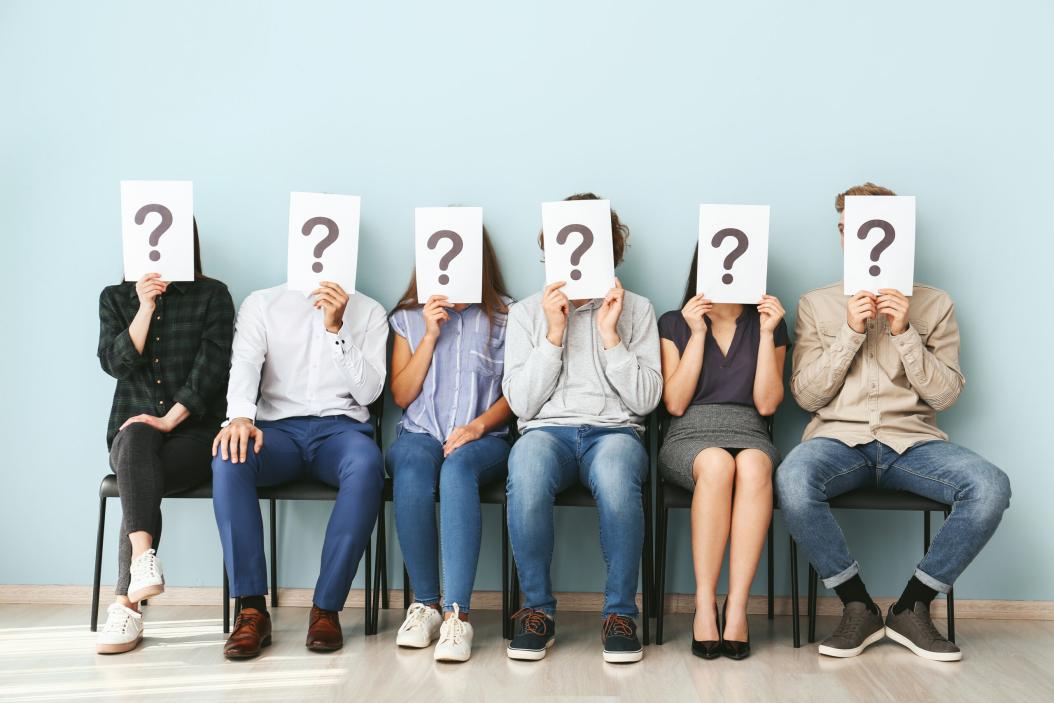 Sechs Personen sitzen auf Stühlen in einer Reihe mit dem Rücken zur Wand. Alle halten vor ihren Gesichtern ein Schild mit einem Fragezeichen darauf.