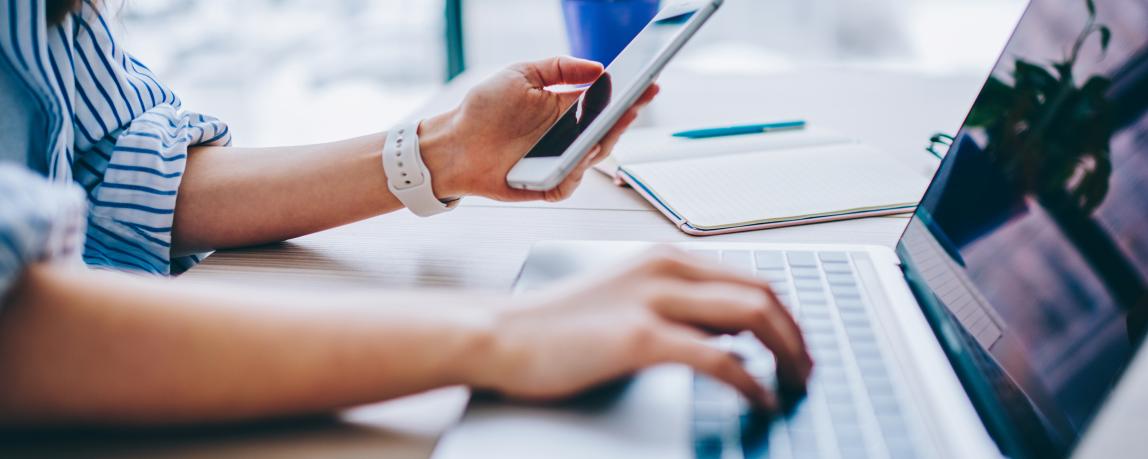 Nahaufnahme einer Frau, die ein Smartphone hält und parallel auf ihrem Laptop tippt