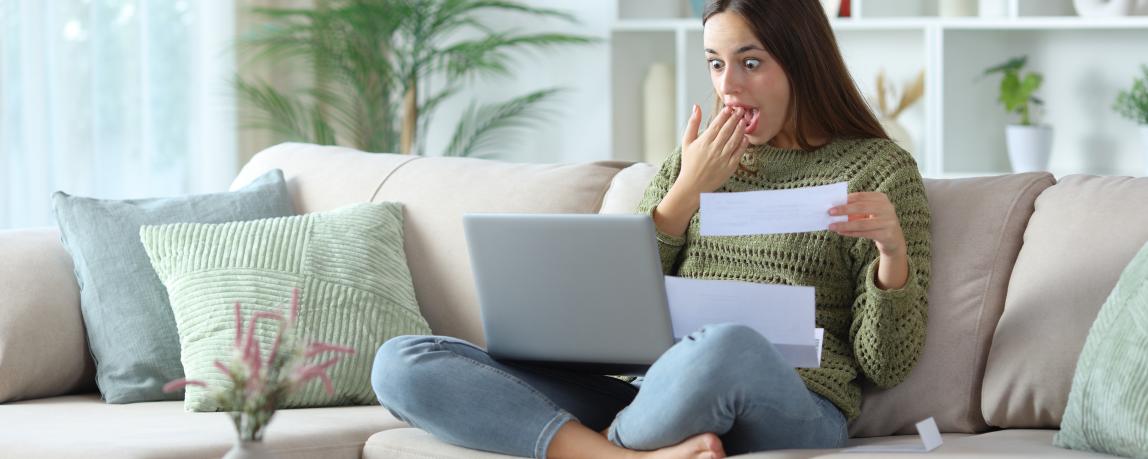 Eine Frau sitzt auf dem Sofa mit dem Laptop auf ihrem Schoß und guckt dabei positiv überrascht.