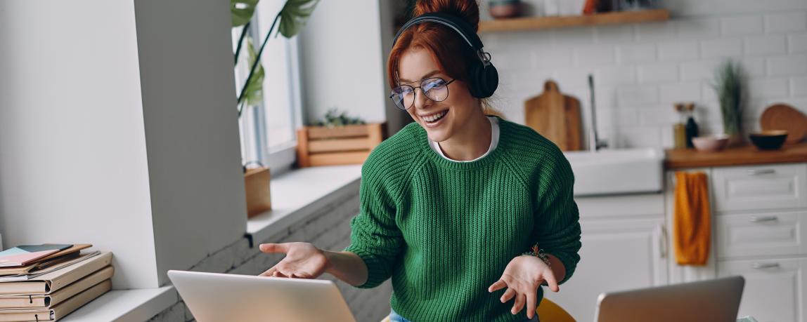 Fröhliche junge Frau mit Kopfhörern nimmt während der Arbeit im Homeoffice an einer Webkonferenz teil.