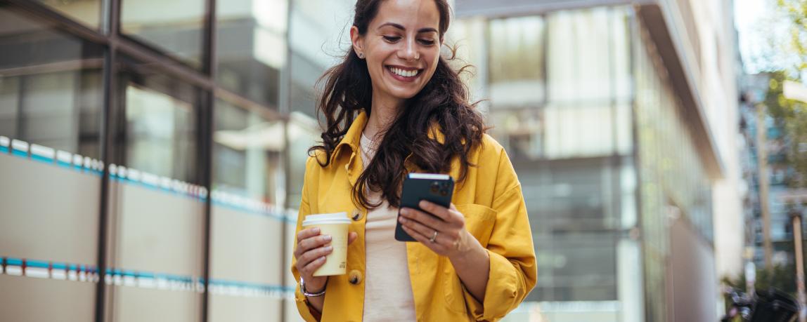 Eine junge Frau geht eine Straße in der Stadt entlang, schreibt Nachrichten und hält dabei einen Kaffeebecher in der Hand.