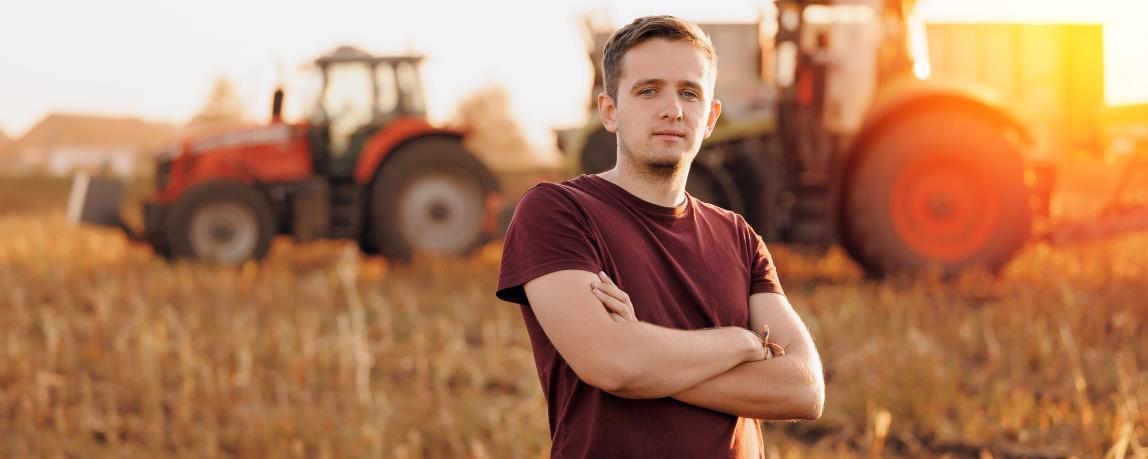 Porträt eines jungen Landwirts in einem Maisfeld, wo im Hintergrund ein Mähdrescher und ein Traktor auf dem abgeernteten Maisfeld zu sehen sind